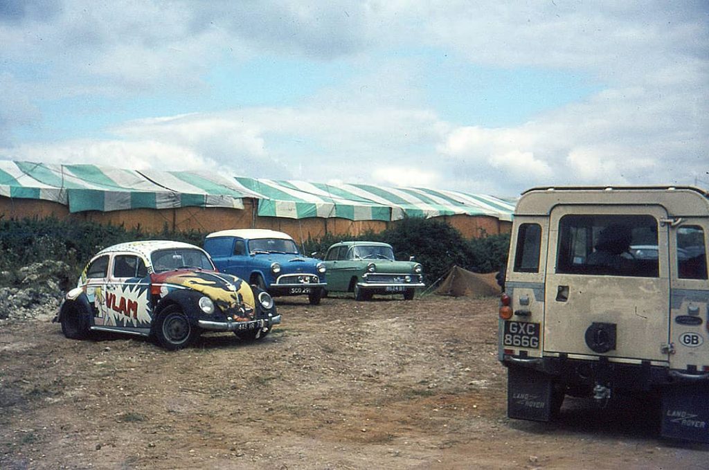 Photograph of multiple cars behind a big communal tent, including what it looks like a Volkswagen Beetle with different drawings on it at the 'Isle of Wight Festival' (1969)