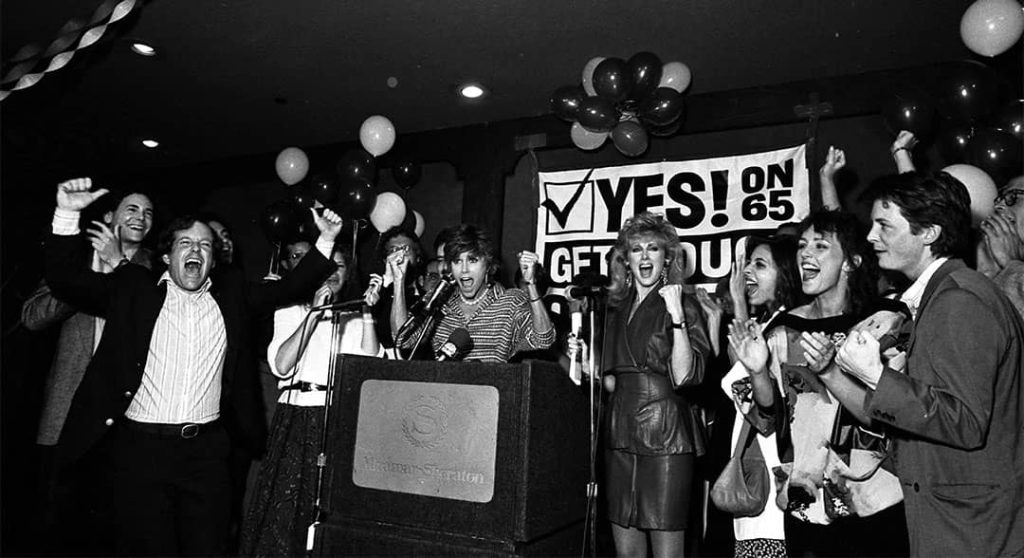 Jane Fonda (centre, at the podium), Morgan Fairchild (to Fonda's right) and Michael J. Fox (far right) along with other people cheering for Proposition 65 at Miramar Sheraton in Santa Monica, California, USA (1986)