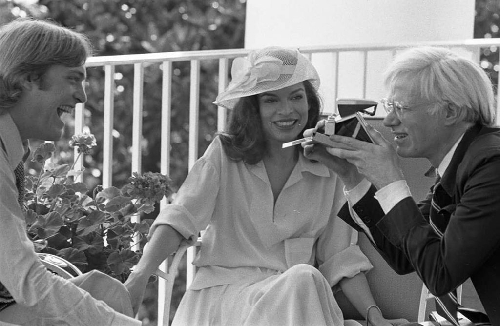 Andy Warhol (right) taking a polaroid picture of Jack Ford (left), Bianca Jagger (centre) is also sitting with them on the Truman Balcony (1975)